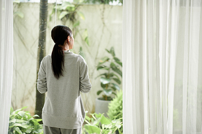 Pensive woman in comfy loungewear looking outside through big window, view from behind