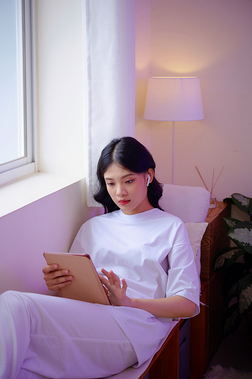 Pensive Asian young woman listening to music in earbuds when playing game on tablet computer