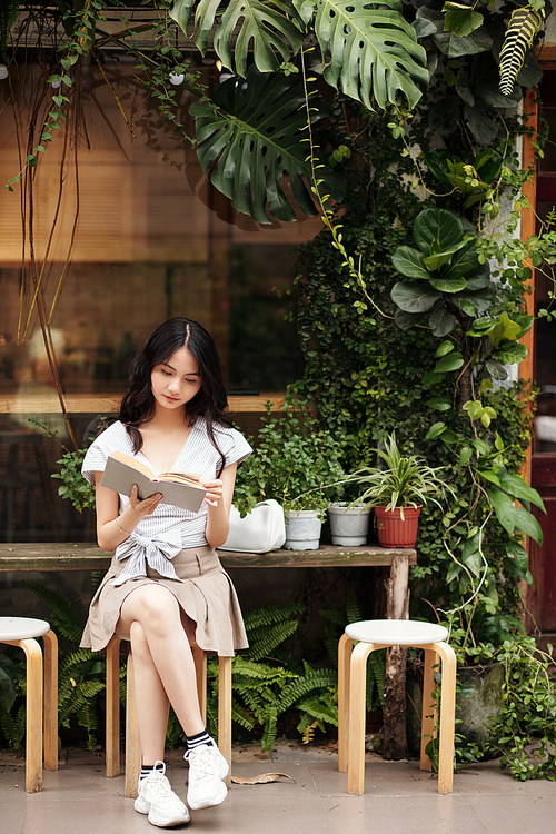Asian young woman sitting outside and reading captivating book