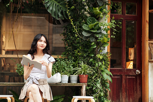 Pensive young Asian woman sitting outside cafe and reading interesting book