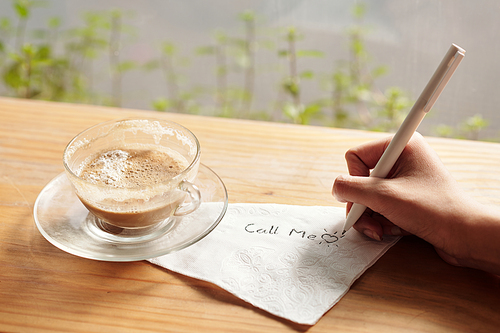 Young woman drinking cup of coffee, writing call me on napkin and drawing heart