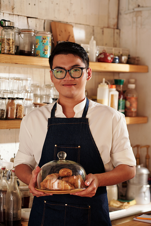 Portrait of smiling young cafe waiter holding plate with hot croissant baked this morning for customers
