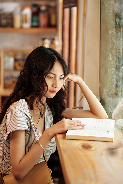 Pensive young Asian woman reading book on cafe