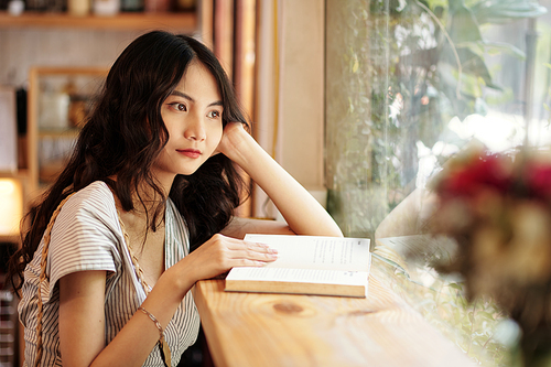 Pensive young woman sitting in cafe with opened book and looking through window
