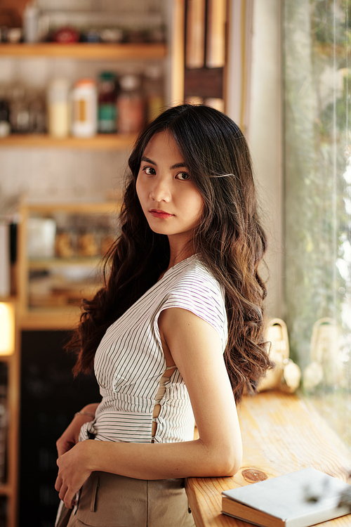 Portrait of serious young Vietnamese woman in striped blouse standing in cafe