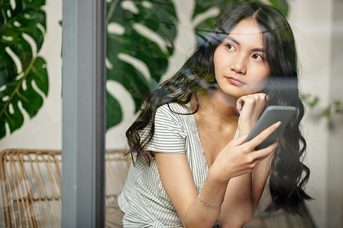 Portrait of pensive young Asian woman with smartphone looking through cafe window