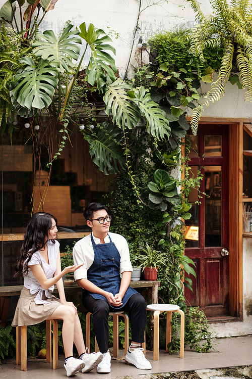 Young barista and his girlfriend sitting outside coffeeshop and waiting for customers