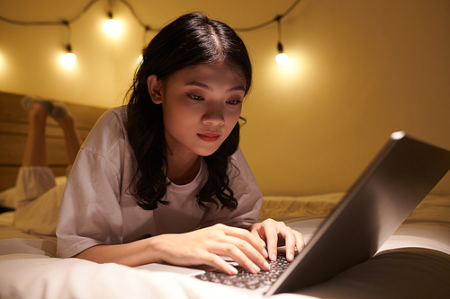 Pensive Vietnamese girl studying late at night when lying on bed with laptop