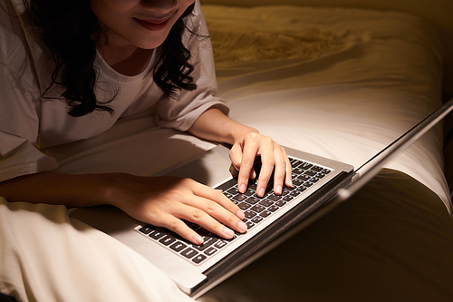 Cropped image of smiling teenage girl working on laptop late at night when lying on bed
