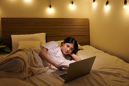 Teenage girl lying on bed under throw blanket and watching show on laptop