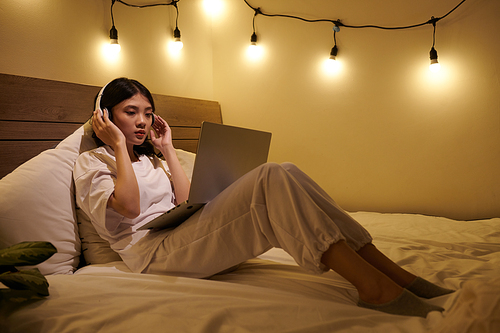 Teenage girl in loungewear in headphones watching webinar on laptop when lying on bed at home