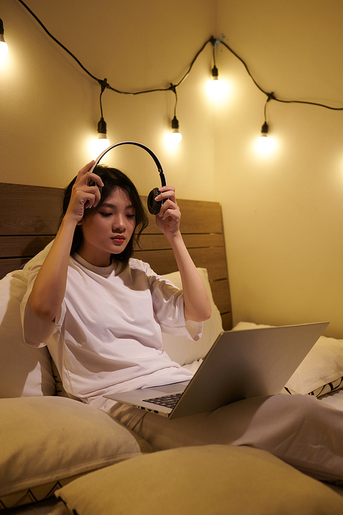 Female gamer putting on headphones to play online game on laptop when sitting in bed