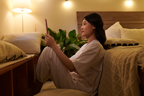 Positive young Asian woman with tablet computer making video call when sitting on floor in bedroom