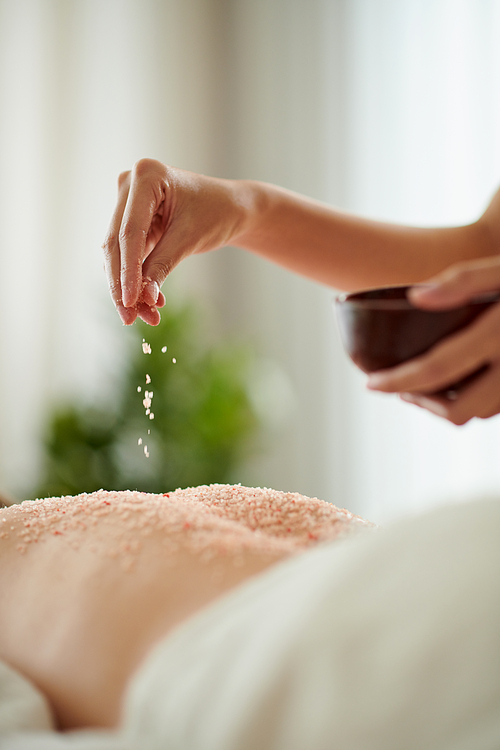 Closeup image of woman pinching infused salt scrub on back of female client of spa salon