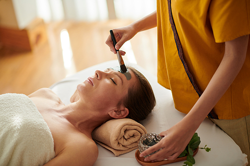 Esthetician applying detoxifying clay mask on face of female client