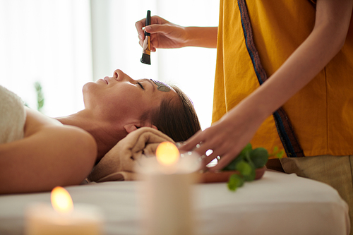 Closeup image of beautician applying detoxifying and brightening clay mask on face of mature woman