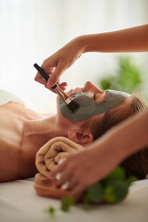 Beautician applying black clay mask on face of client in beauty salon