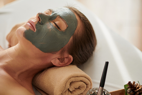 Woman with brightening clay mask on face relaxing on bed in spa salon