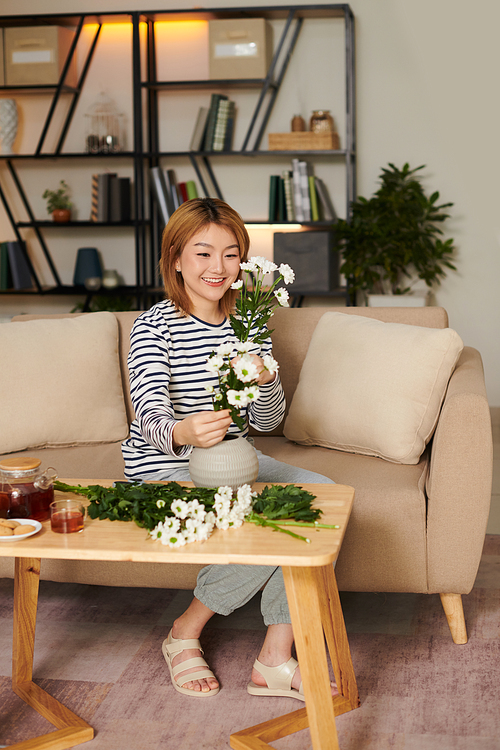 Smiling young Asian woman arranging daisy flowers in bouquet at home
