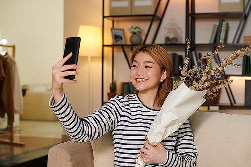 Positive young Asian woman taking selfie with bouquet of dried flowers to post on social media
