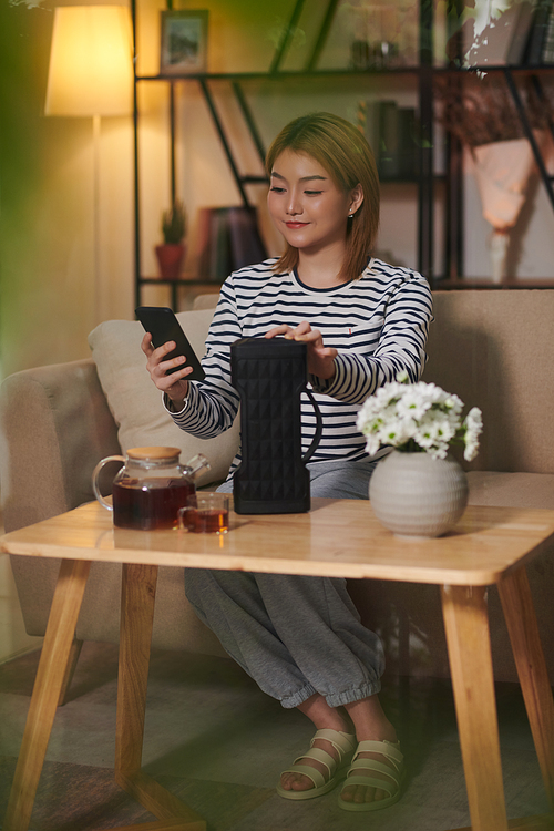 Young Asian woman turning on calm music on wireless speaker to relax when enjoying cup of tea