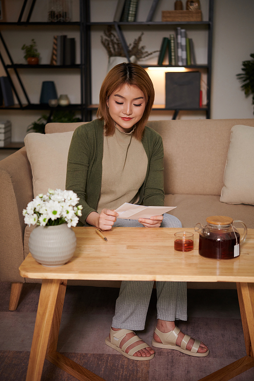 Young Vietnamese woman drinking tea and reading letter from friend when spending evening at home