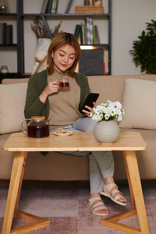Young woman drinking tea with cookies and scrolling social media on smartphone
