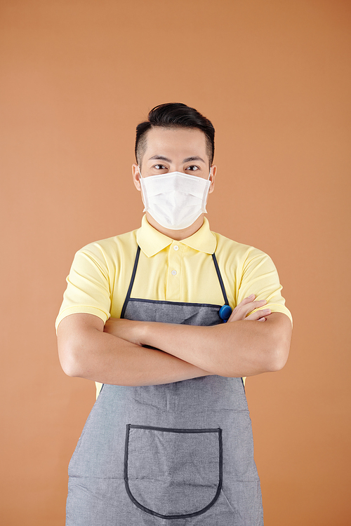 Portrait of coffeeshop barista in apron and medical mask crossing arms and looking at camera