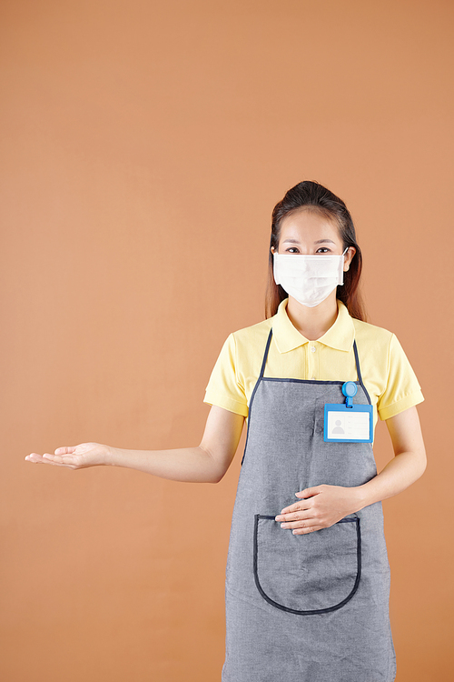 Studio portrait of positive young coffeeshop barista in medical mask protecting against coronavirus inviting customers