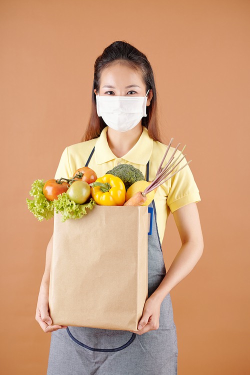 Smiling young supermarket worker in medical mask holding paper bag full of fresh groceries