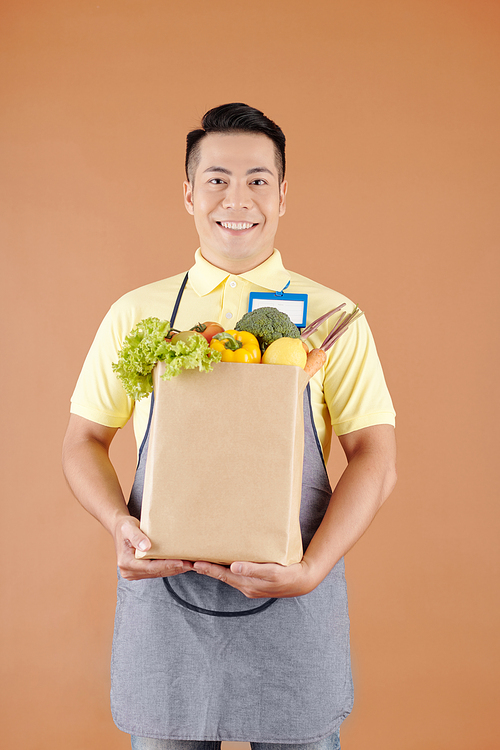 Smiling Asian supermarket worker holding paper bag full of fresh groceries, fruits and vegetables