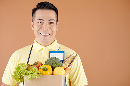 Portrait of cheerful delivery service or dark store worker holding paper bag of fresh vegetables