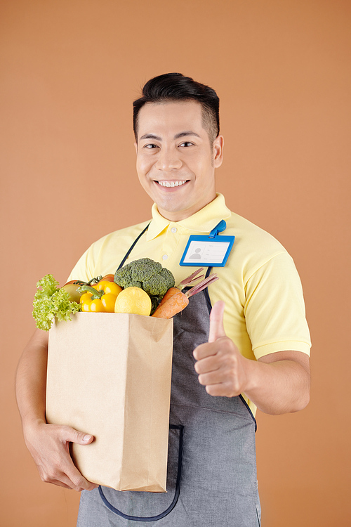 Portrait of happy supermarket worker holding paper bag with groceries and showing thumbs-up
