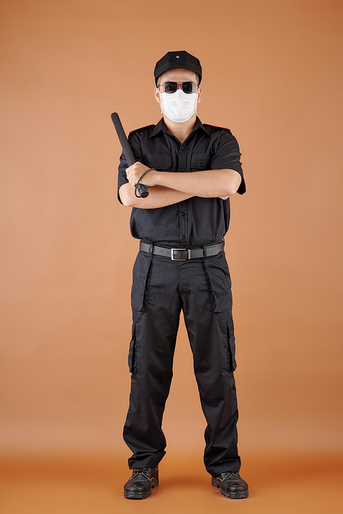 Portrait of confident security guard in medical mask holding baton, crossing arms and looking at camera