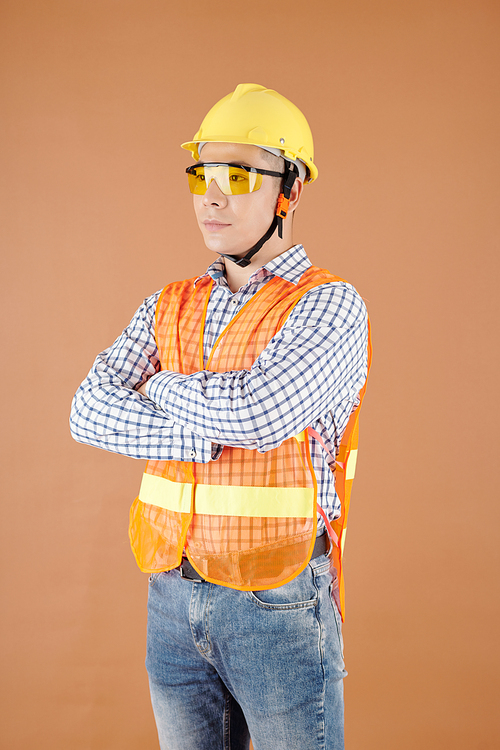 Portrait of serious construction worker crossing arms and looking away, isolated on brown