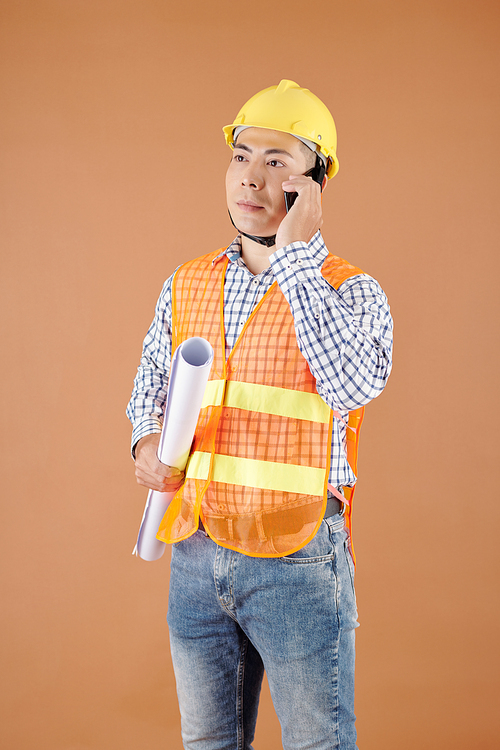 Portrait of serious construction engineer with blueprint in hand talking on phone with contractor
