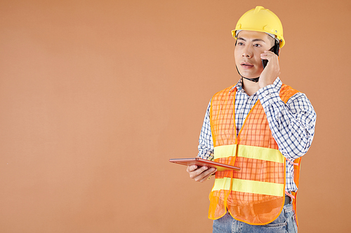 Serious construction engineer holding tablet computer when talking on phone with foreman or contractor