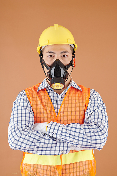 Studio portrait of workman in hardhat and breathing mask crossing arms and looking at camera