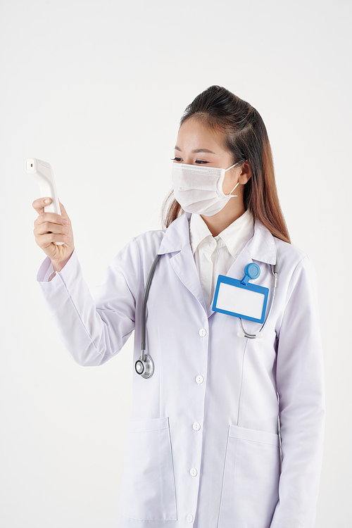 Young Asian doctor in medical mask checking body temperature on electronic contactless thermometer