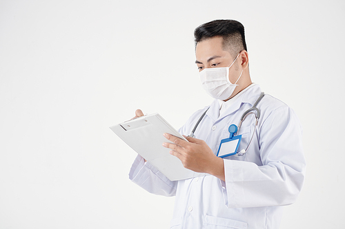 Studio portrait of doctor in protective mask filling medical report on clipboard