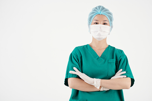 Portrait of female surgeon in scrubs, gloves, hat and medical mask crossing arms and looking at camera