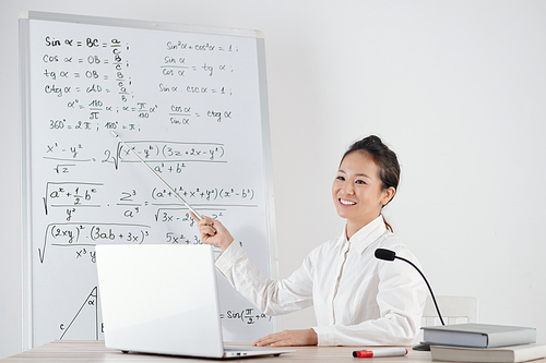 Smiling young Asian mathematics teacher pointing at whiteboard with formulas when explaining topic online to students