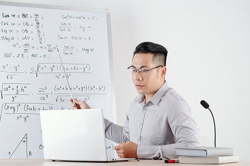 Asian math teacher pointing at whiteboard with formulas when explaining topic to college students during online class