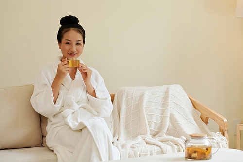 Portrait of happly young Asian woman resting on couch after taking bath and drinking herbal tea