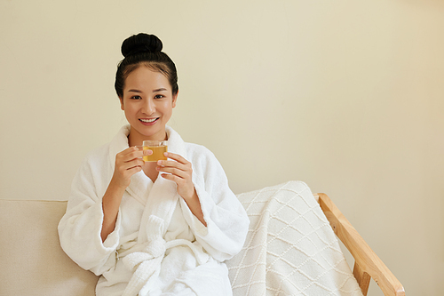 Portrait of smiling young Vietnamese woman in bathrobe holding cup of herbal tea and looking at camera