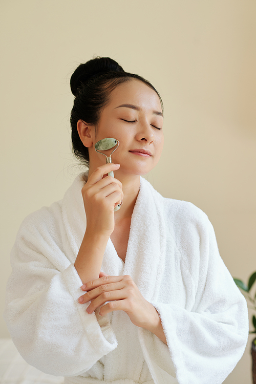 Young woman closing eyes when massaging face with jade roller after applying moisturizing serum