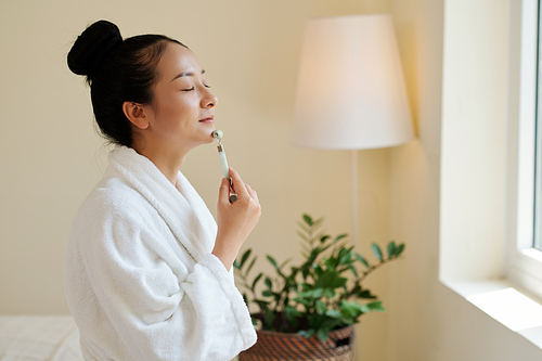 Smiling young woman in bathrobe massaging face with v-shape roller to stimulate blood flow and make skin look brighter
