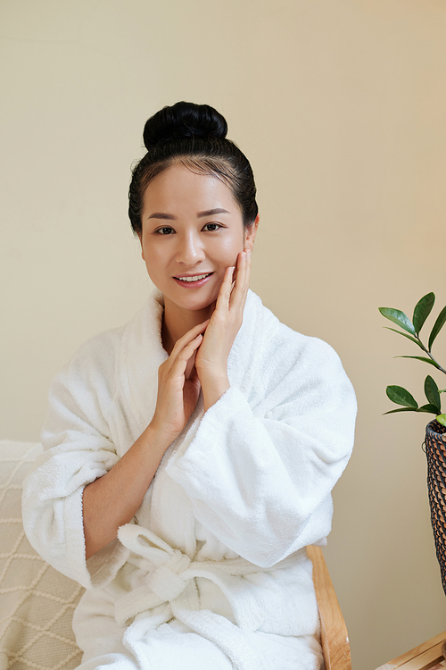 Portrait of smiling young woman in bathrobe touching soft skin of her face after applying moisturizing lotion