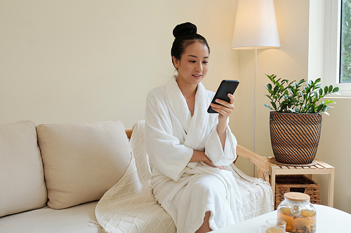 Positive young Vietnamese woman in bathrobe resting on couch after spa procedure and reading news on social media