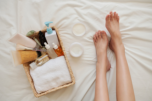 Feet of young woman on bed next to basket of skincare products like cream, lotions and oils